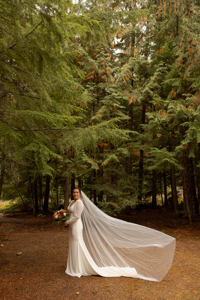 A bride poses with her bouquet and her veil flowing in the wind in a forest at her Idaho elopement