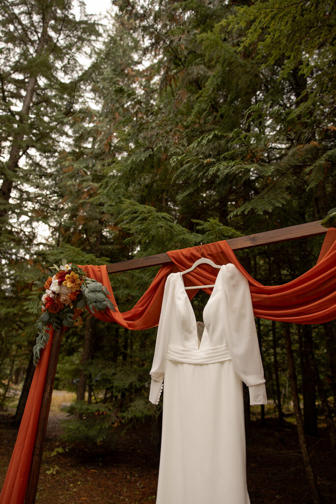 Priest Lake Idaho Wedding | Love In The Hills Photo And Video Wedding dress hanging on a wooden arbor surrounded by towering trees before a Priest Lake Idaho wedding