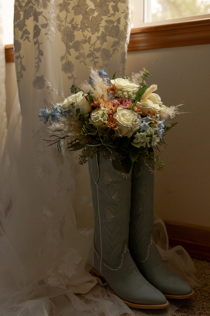Priest Lake Wedding | Love In The Hills Photo And Video Blue cowboy boots filled with a bridal bouquet beside a wedding dress for a Priest Lake Idaho wedding.
