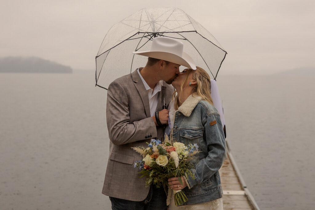 Priest Lake Idaho Wedding | Love In The Hills Photo And Video Bride and groom kissing beneath a clear umbrella on a dock during a Priest Lake Idaho wedding.